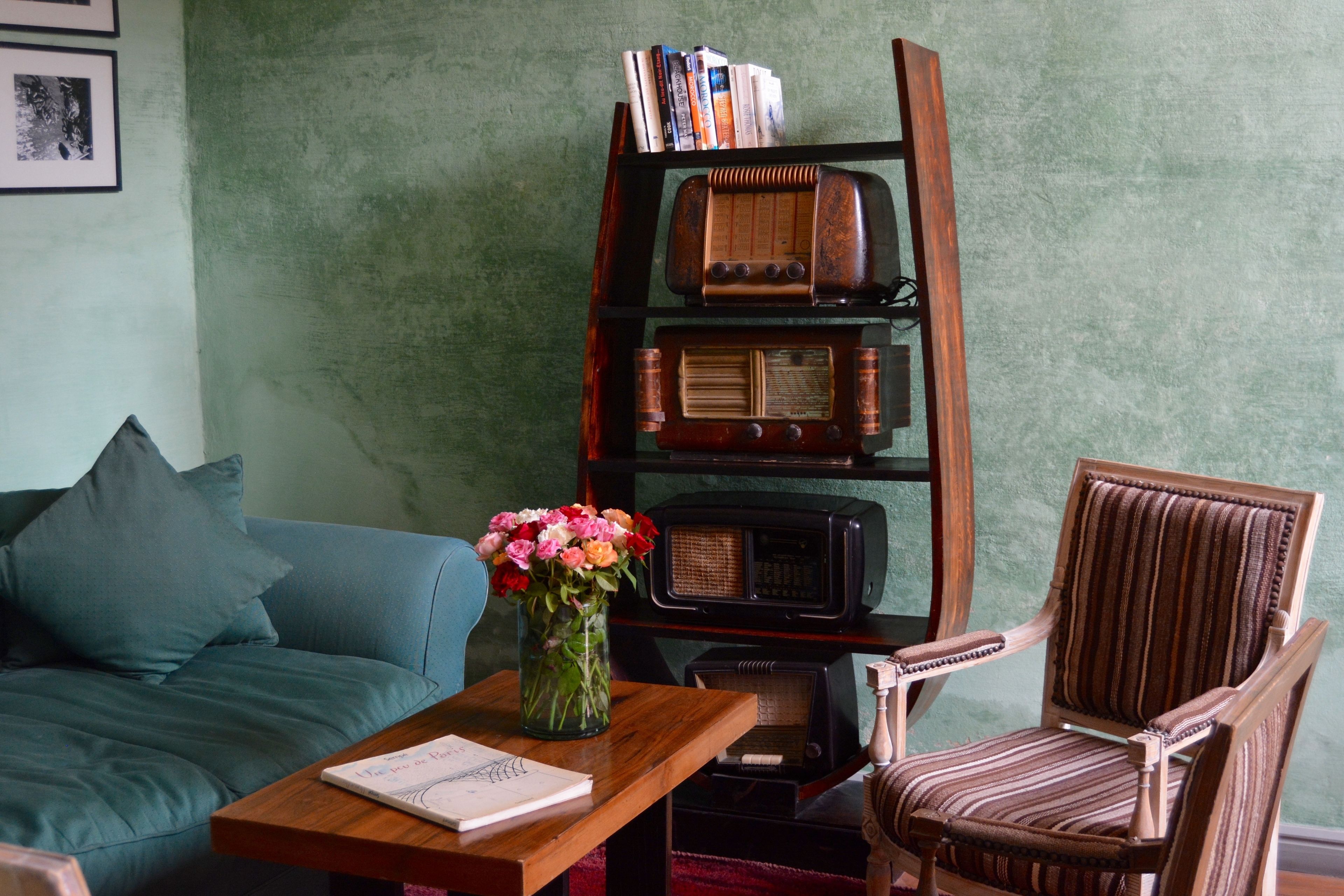 Una sala de estar con un sofá azul, una mesa de centro de madera con un libro y un jarrón de flores, una estantería de madera con libros y una radio antigua, y un sillón a rayas.