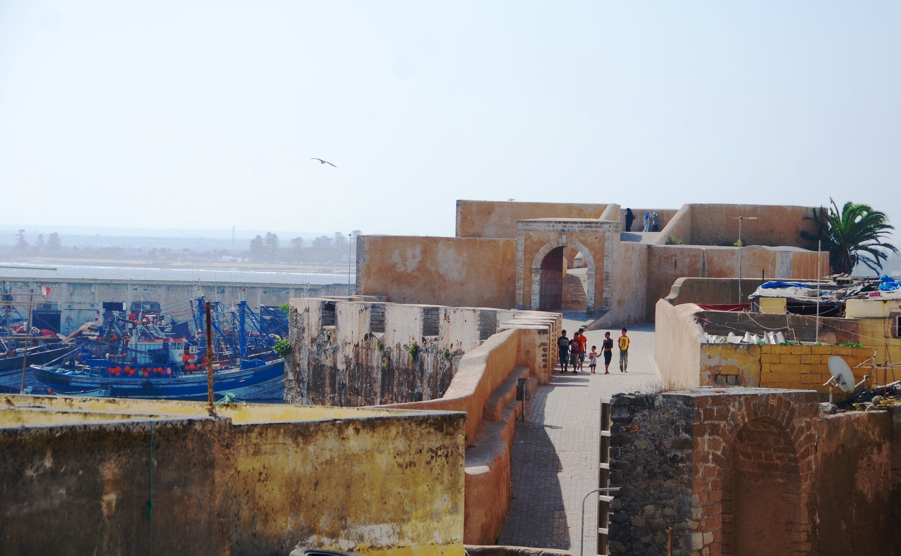 Una zona costera con barcos amarrados, una estructura fortificada y un sendero con gente caminando.