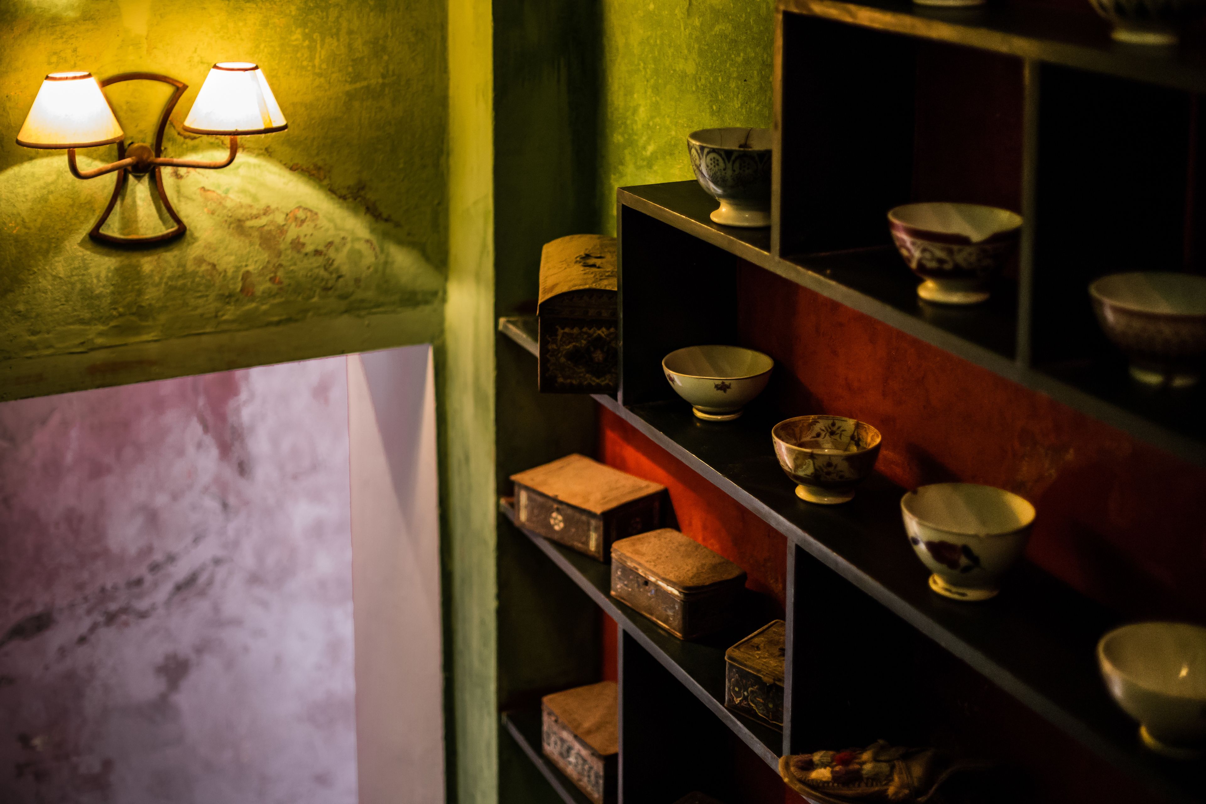 Una habitación con poca luz, paredes verdes, una estantería con cuencos y cajas varios, y una lámpara fijada a la pared.