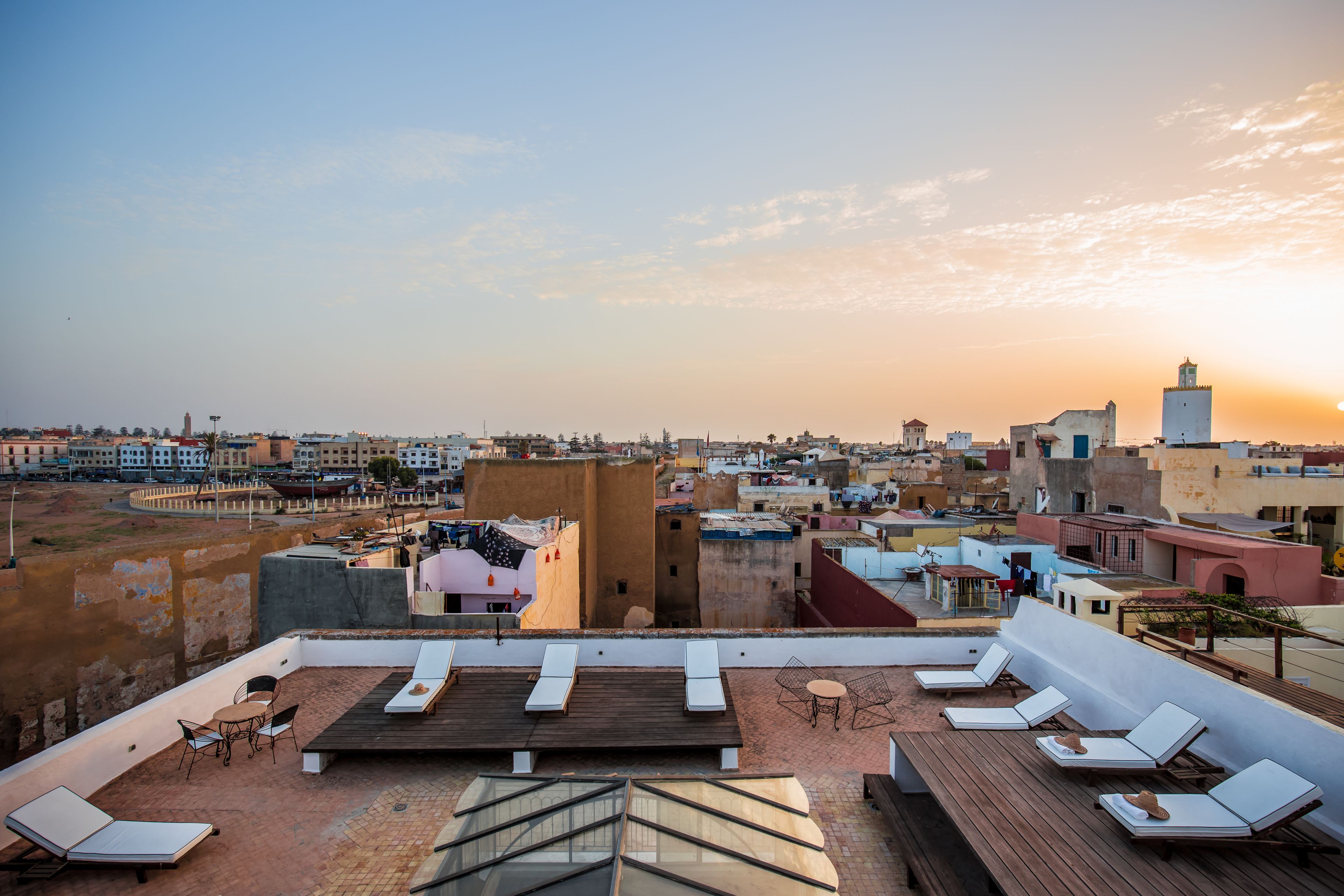 Terraza en la azotea con tumbonas y una mesa, con vistas a un paisaje urbano con edificios y una torre.