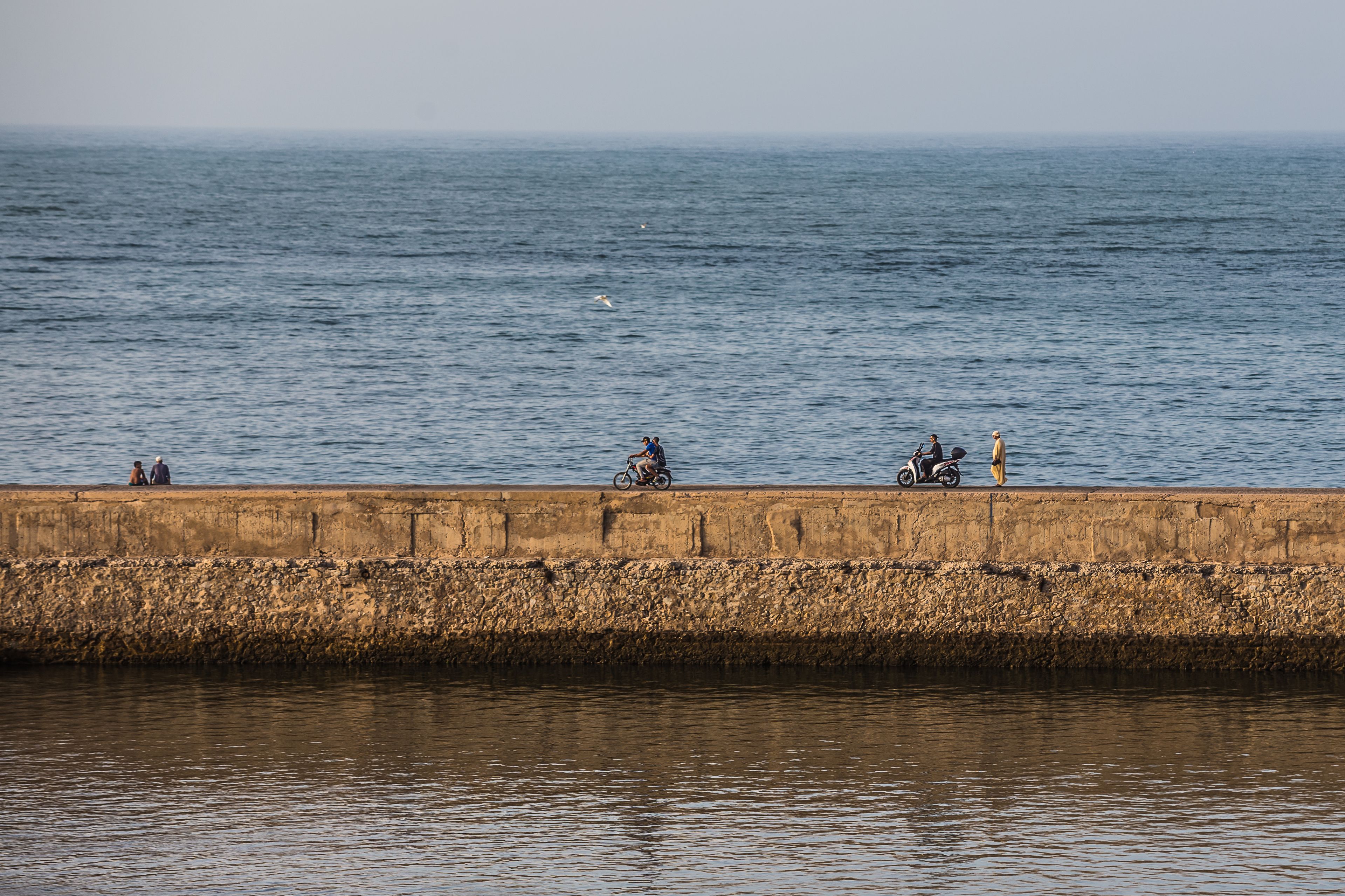 Une vue sur le bord de l’eau, avec une barrière en béton, une eau calme et quelques personnes et scooters le long du quai.