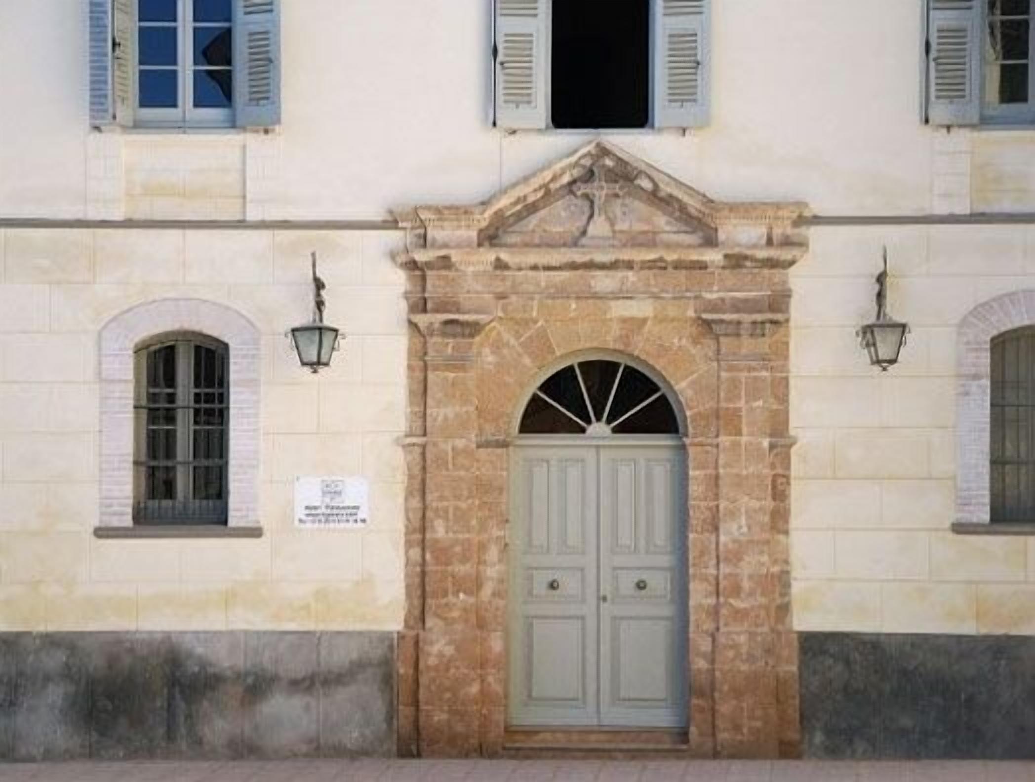 Fachada de edificio con una entrada arqueada central, flanqueada por dos ventanas con contraventanas y dos faroles de pared.