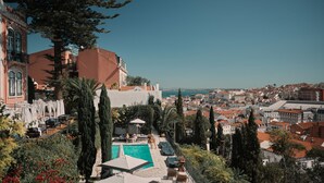 Un área de piscina en la azotea con sillones de descanso y sombrillas, rodeada de árboles y con vistas a una ciudad costera.