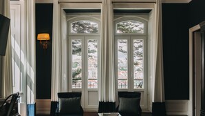 Una habitación con dos sillones negros, una mesita y una lámpara. Amplias ventanas con cortinas blancas ofrecen vistas de edificios exteriores.