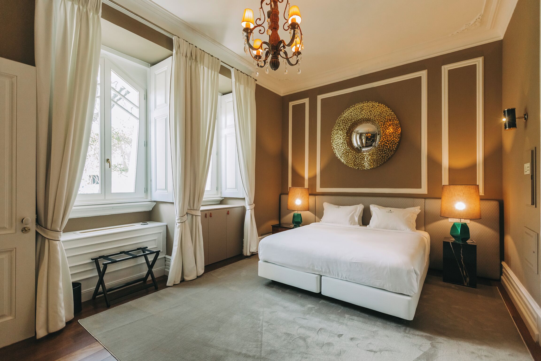 Ein Schlafzimmer mit einem großen Bett, zwei Nachttischlampen, einer Chandelüster, einem runden Spiegel an der Wand und einem Fenster mit Vorhängen.