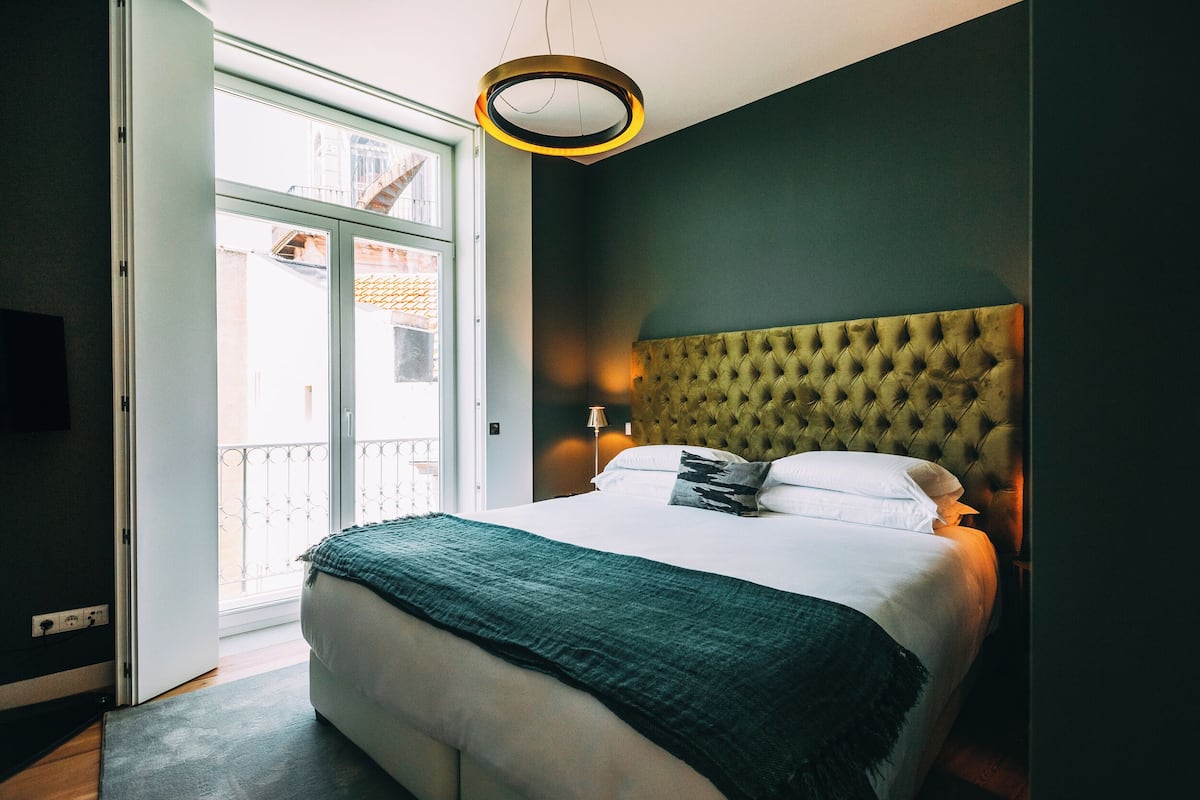 Une chambre à coucher avec un lit, une tête de lit, une lampe de chevet et une vue sur l’extérieur, visible à travers une porte ouverte.