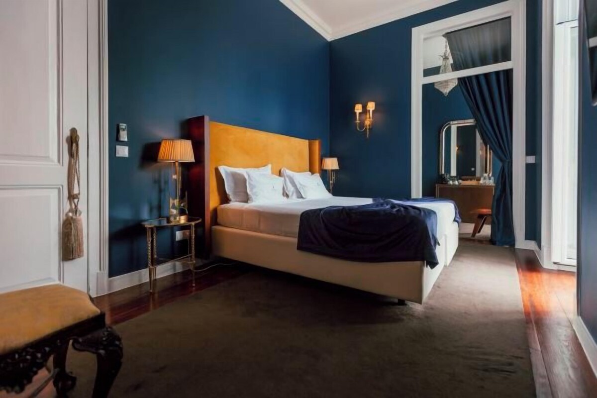 A bedroom with a bed, bedside lamps, a nightstand, a bench, and a window with curtains.