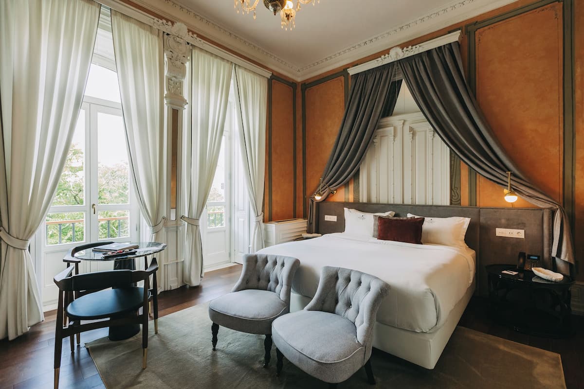 Une chambre d’hôtel avec un grand lit, deux fauteuils, une petite table et une vue sur la verdure.