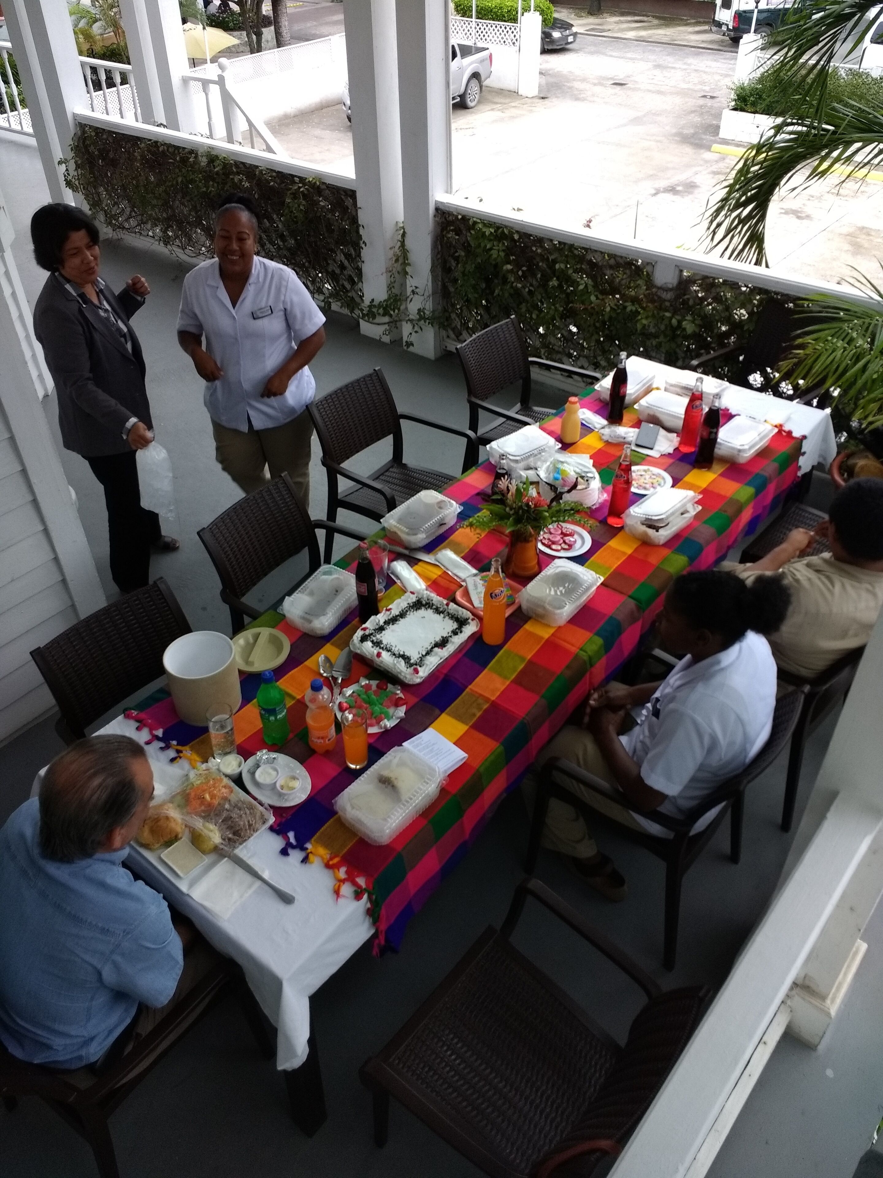 A long dining table with a colorful tablecloth, surrounded by chairs, set for a meal.