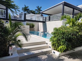 Una piscina con escalones que llevan a una zona cubierta, rodeada de vegetación y una valla blanca.