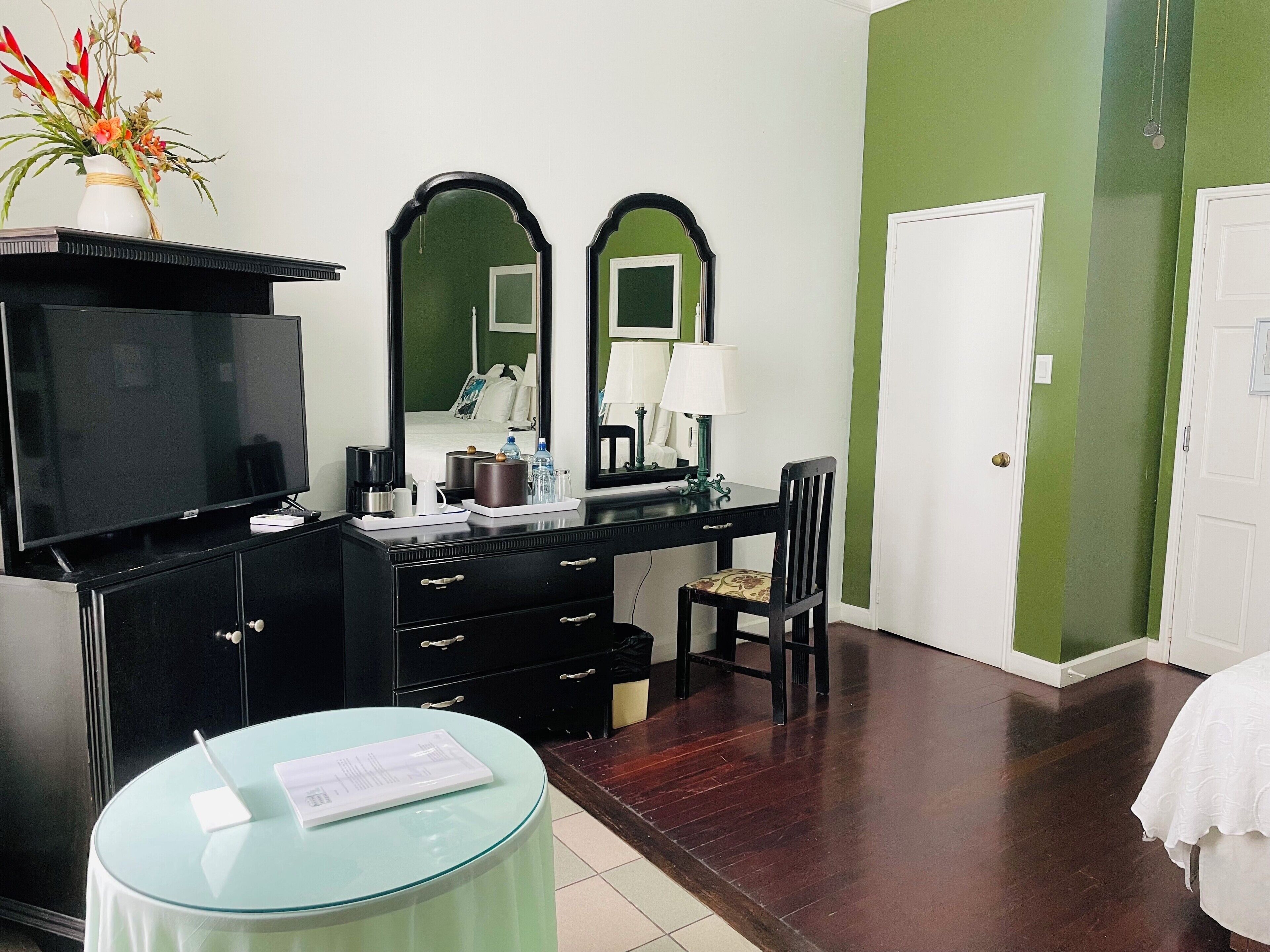 A hotel room with a TV, a desk with a mirror, a chair, a small table with a menu, and a vase with flowers.