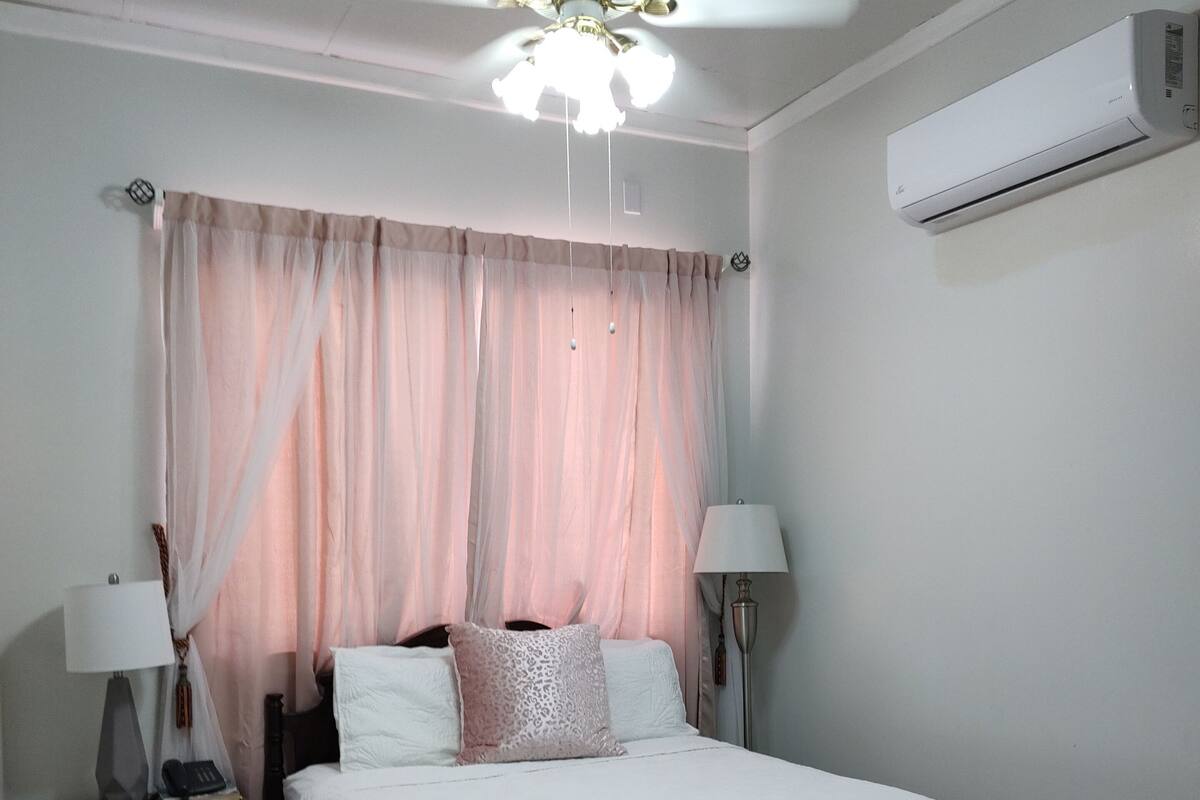 A bedroom with a bed, two bedside tables with lamps, a ceiling fan, and an air conditioning unit.