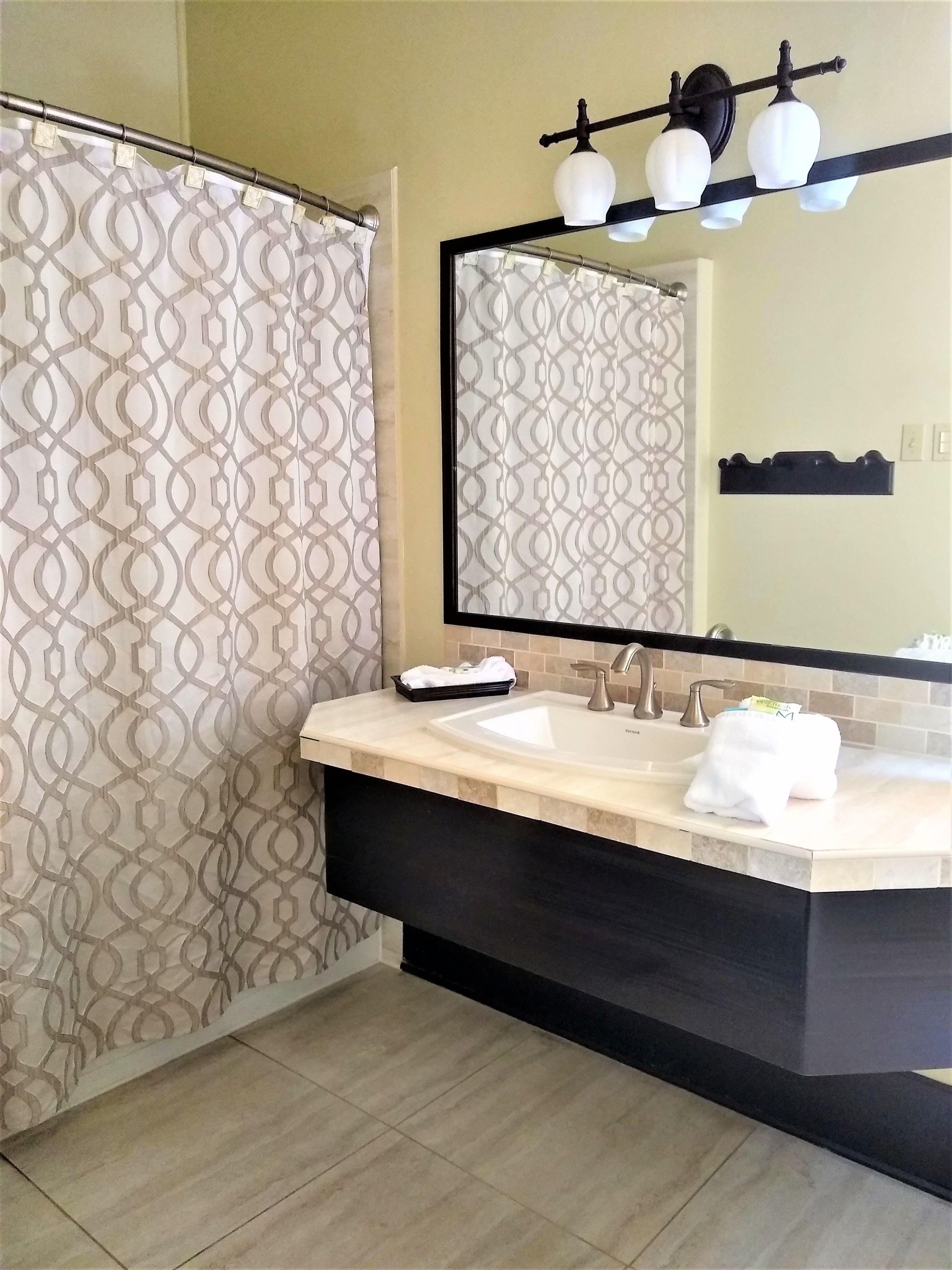 A bathroom with a double sink vanity, a mirror with three lights, and a shower curtain with a geometric pattern.