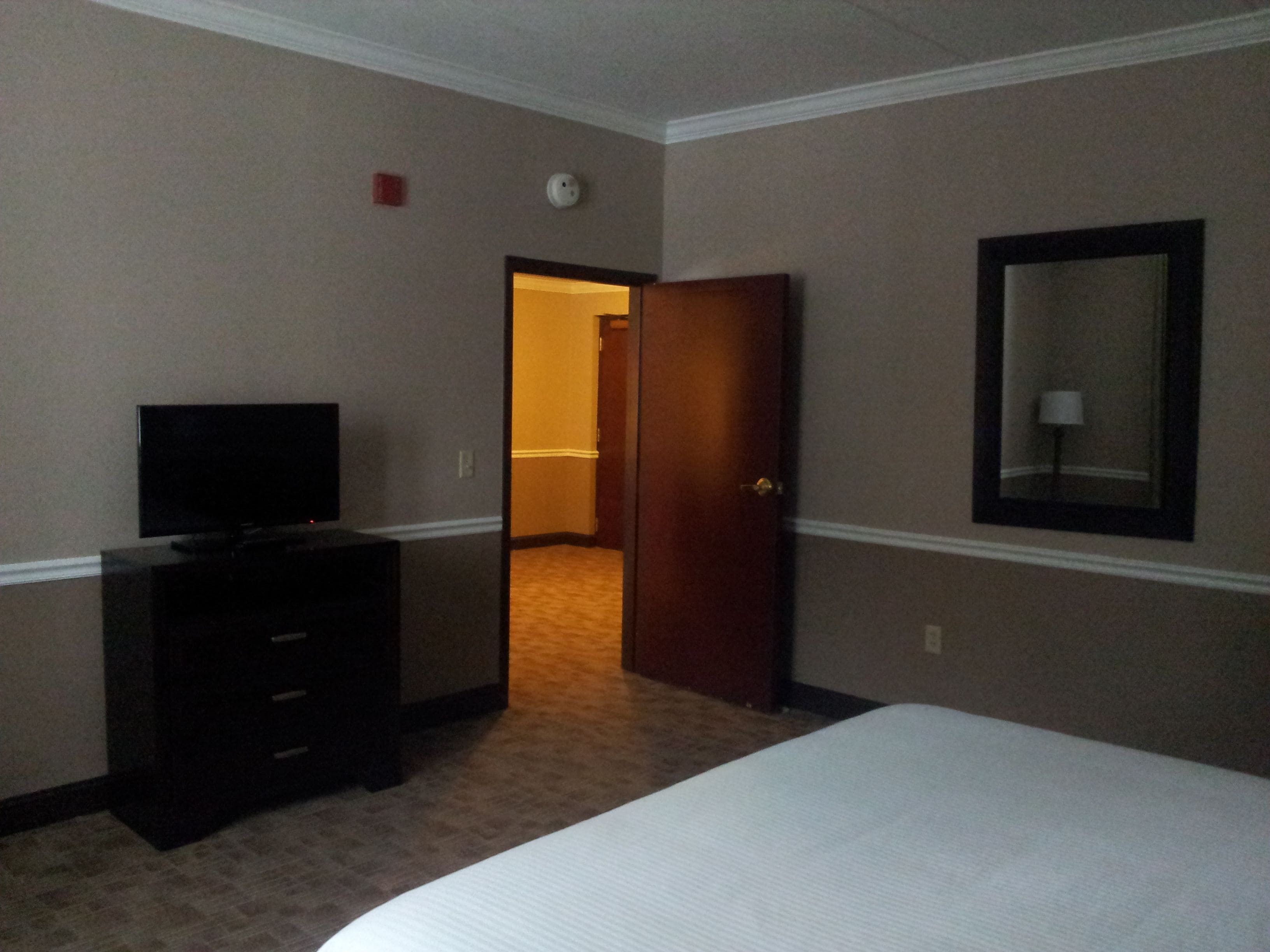 A hotel room with a bed, a dresser, a television, a door leading to another room, and a wall-mounted mirror.