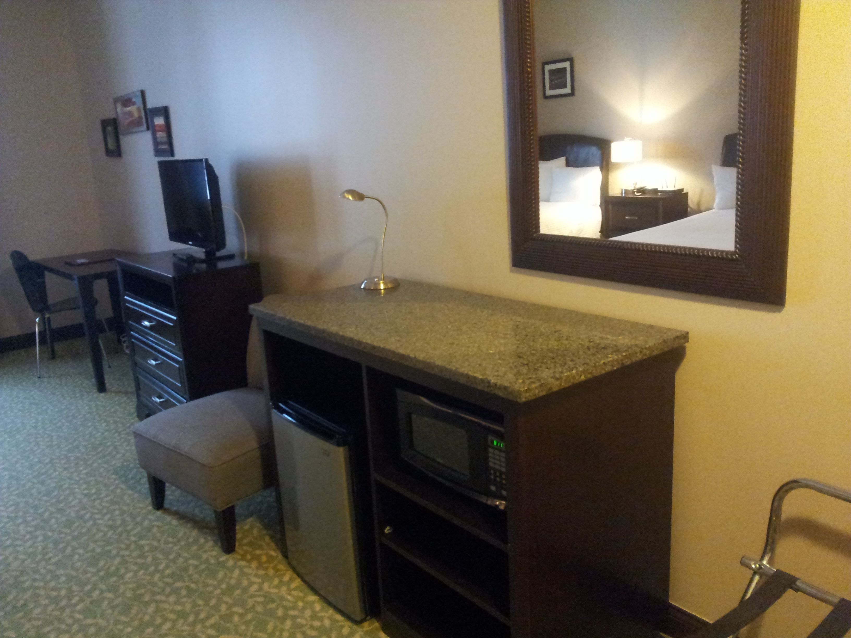 Een compacte hotelkamer met kitchenette, bureau, stoel, kleine koelkast, magnetron en spiegel.