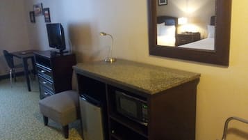 A compact hotel room with a kitchenette, a desk, a chair, a small refrigerator, a microwave, and a mirror on the wall.