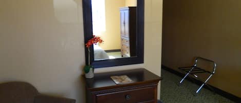 A hotel room with a wooden dresser, a mirror, a small vase with flowers, a chair, and a metal rack.