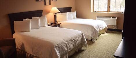 A hotel room with two beds, a nightstand, a lamp, a radiator, and a window with blinds.