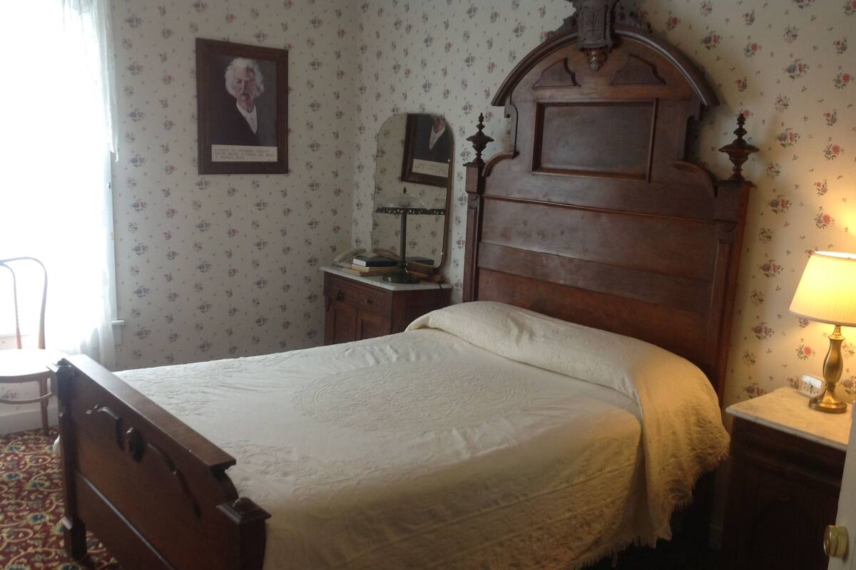 A traditional bedroom with a wooden bed, a nightstand with a lamp, a mirror, and a framed picture on the wall.