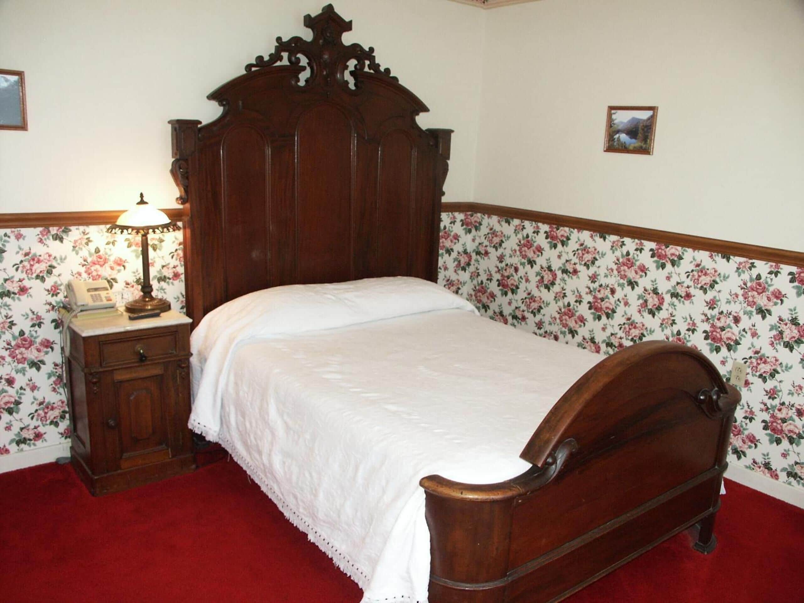 Un dormitorio con una cama de madera, una mesita de noche con lámpara, papel tapiz floral y una alfombra roja.