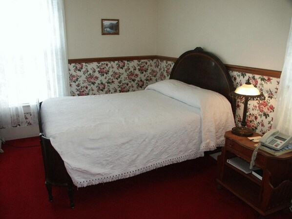 A neatly made bed with a white bedspread and a wooden headboard. There is a wooden nightstand with a lamp and a telephone beside the bed. The room has floral wallpaper and a red carpet. - Bridgeport Inn (Bridgeport)