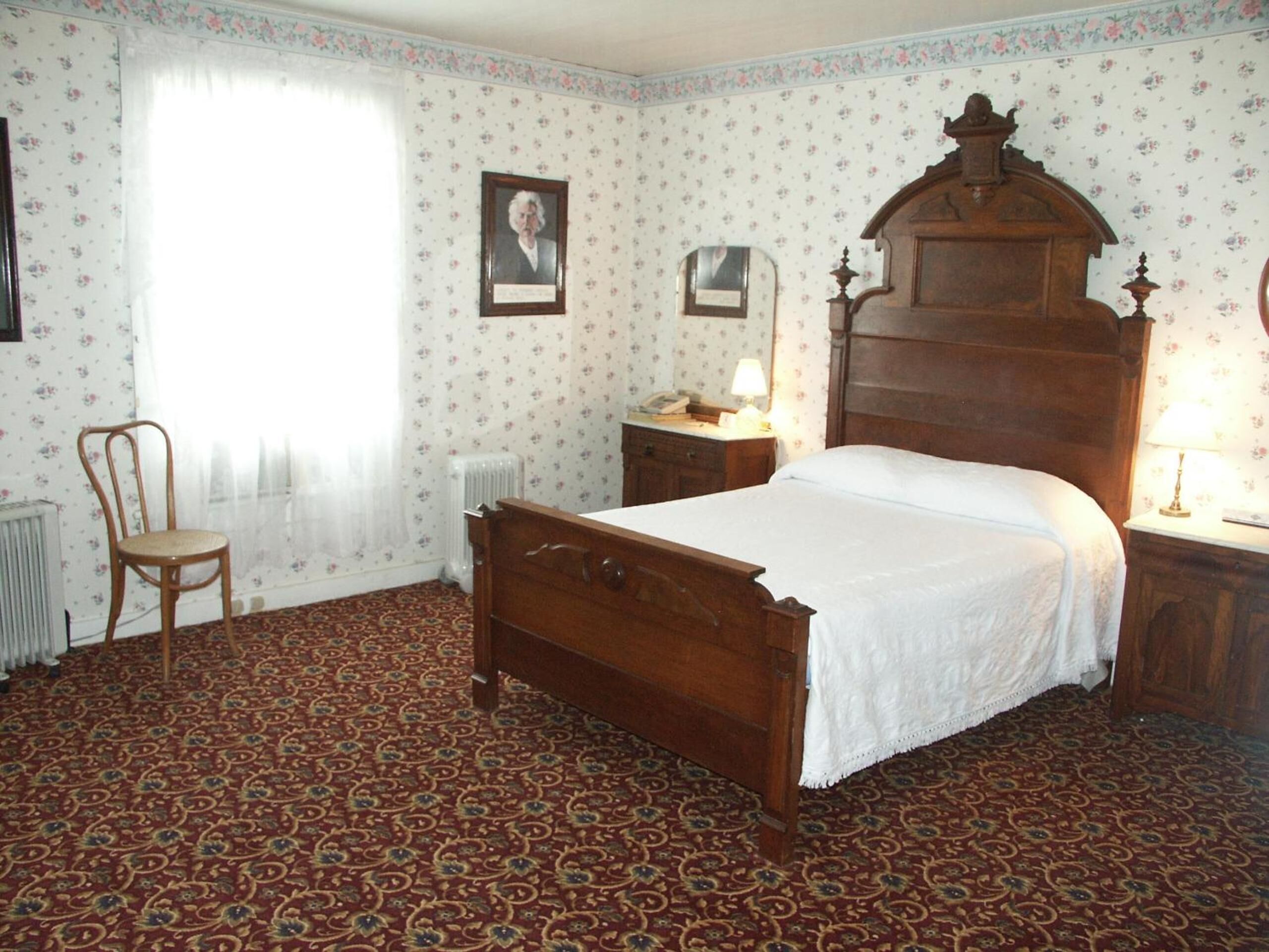A bedroom with a wooden bed, a chair, a nightstand, a radiator, and floral wallpaper.