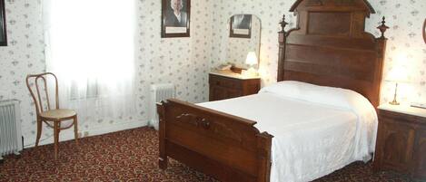 A bedroom with a wooden bed, a chair, a nightstand, a radiator, and floral wallpaper.