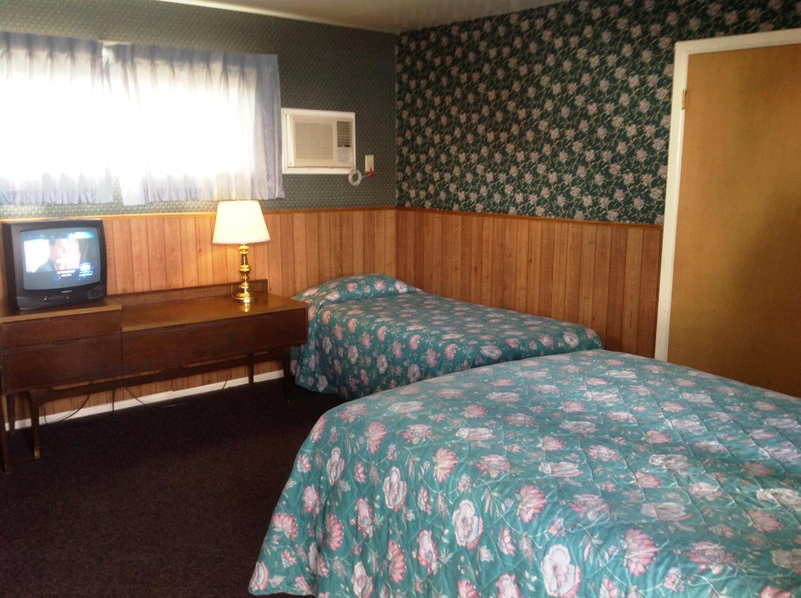 Une chambre avec deux lits, un bureau avec une lampe, une télévision et une fenêtre avec des rideaux.