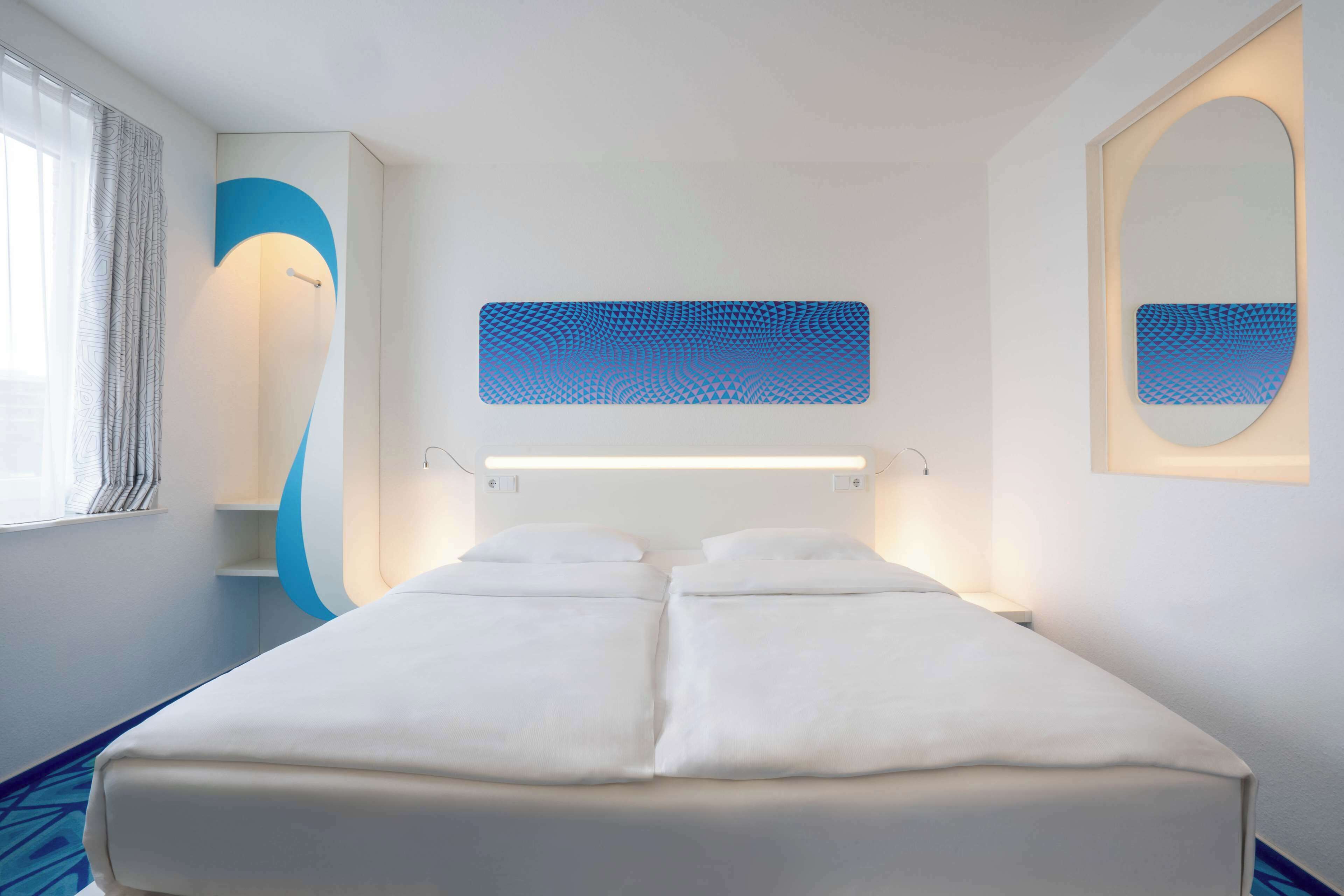 Une chambre d’hôtel moderne avec un grand lit, une table de chevet, un miroir et un luminaire fixé au mur.