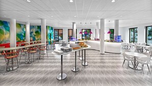 Een modern café met witte meubels, een centrale bedieningsbalie en kleurrijke abstracte muurkunst.