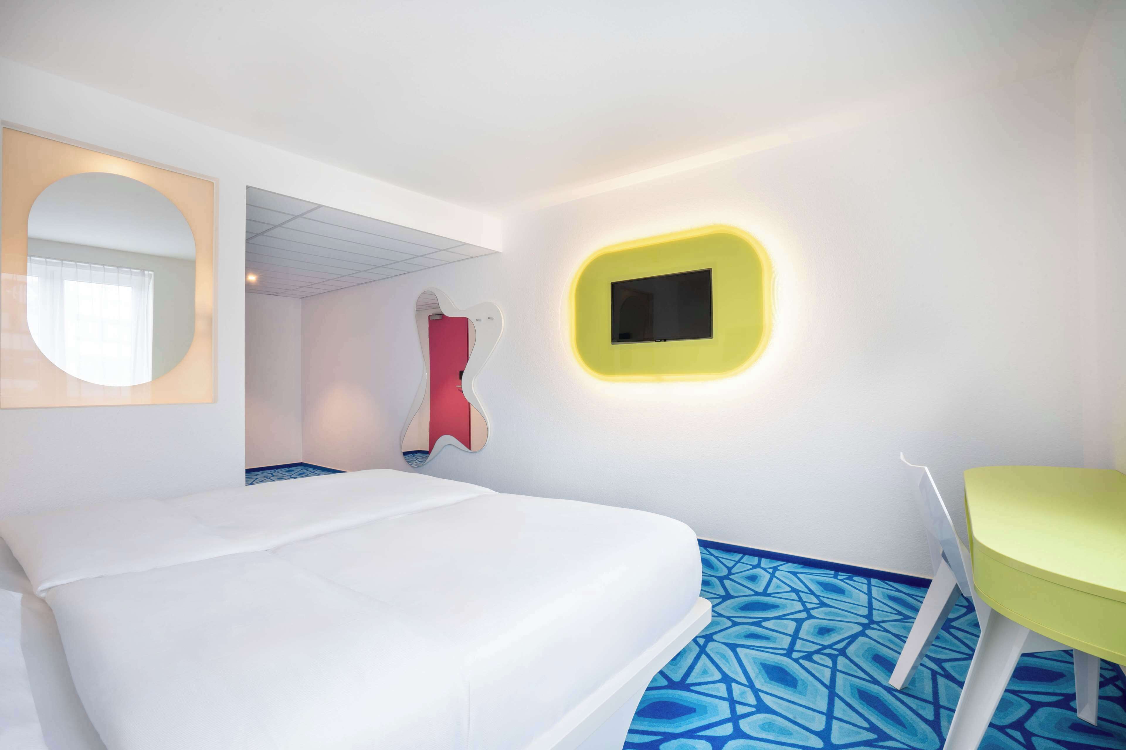 Une chambre d’hôtel avec un lit, un fauteuil vert, un fauteuil rouge, un téléviseur jaune fixé au mur et un sol à motifs.