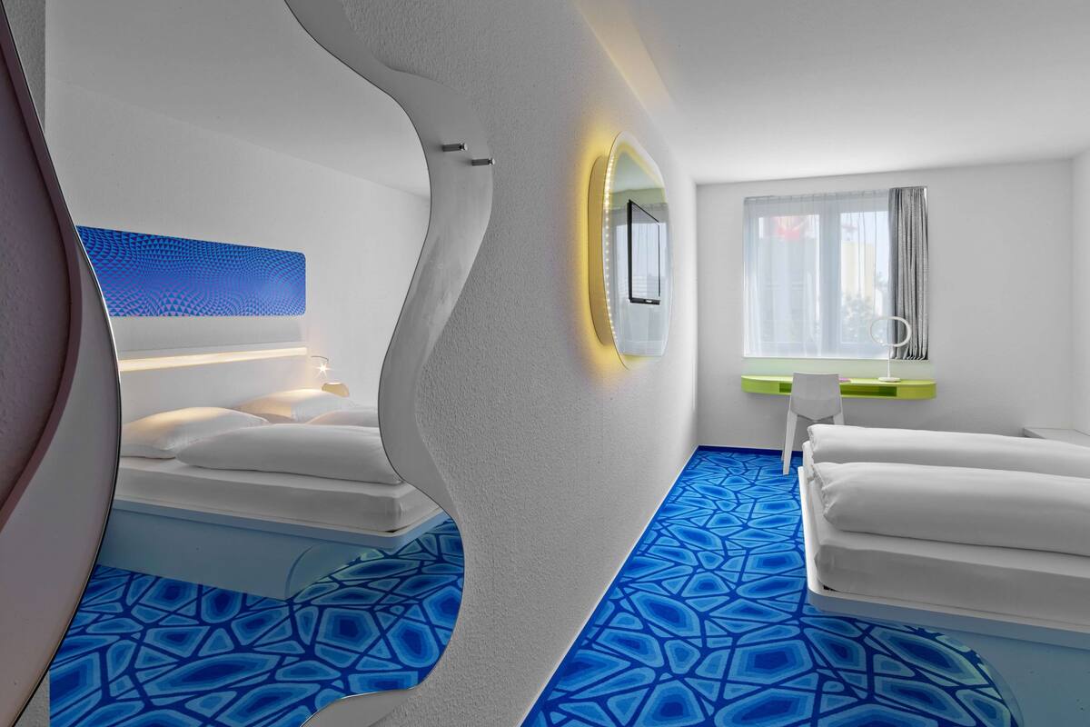 A modern hotel room with a unique curved wall design, a large mirror, a bedside table, a window with curtains, and a patterned blue carpet.