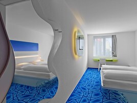 Ein modernes Hotelzimmer mit einem einzigartigen, geschwungenen Wanddesign, einem großen Spiegel, einem Nachttisch, einem Fenster mit Vorhängen und einem gemusterten, blauen Teppich.