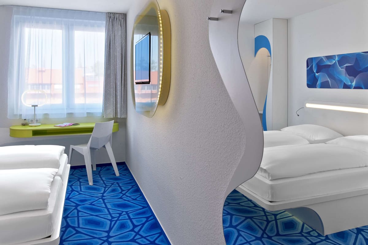 A modern hotel room with a curved wall design, a large mirror, a desk with a chair, two beds, and a patterned blue carpet.
