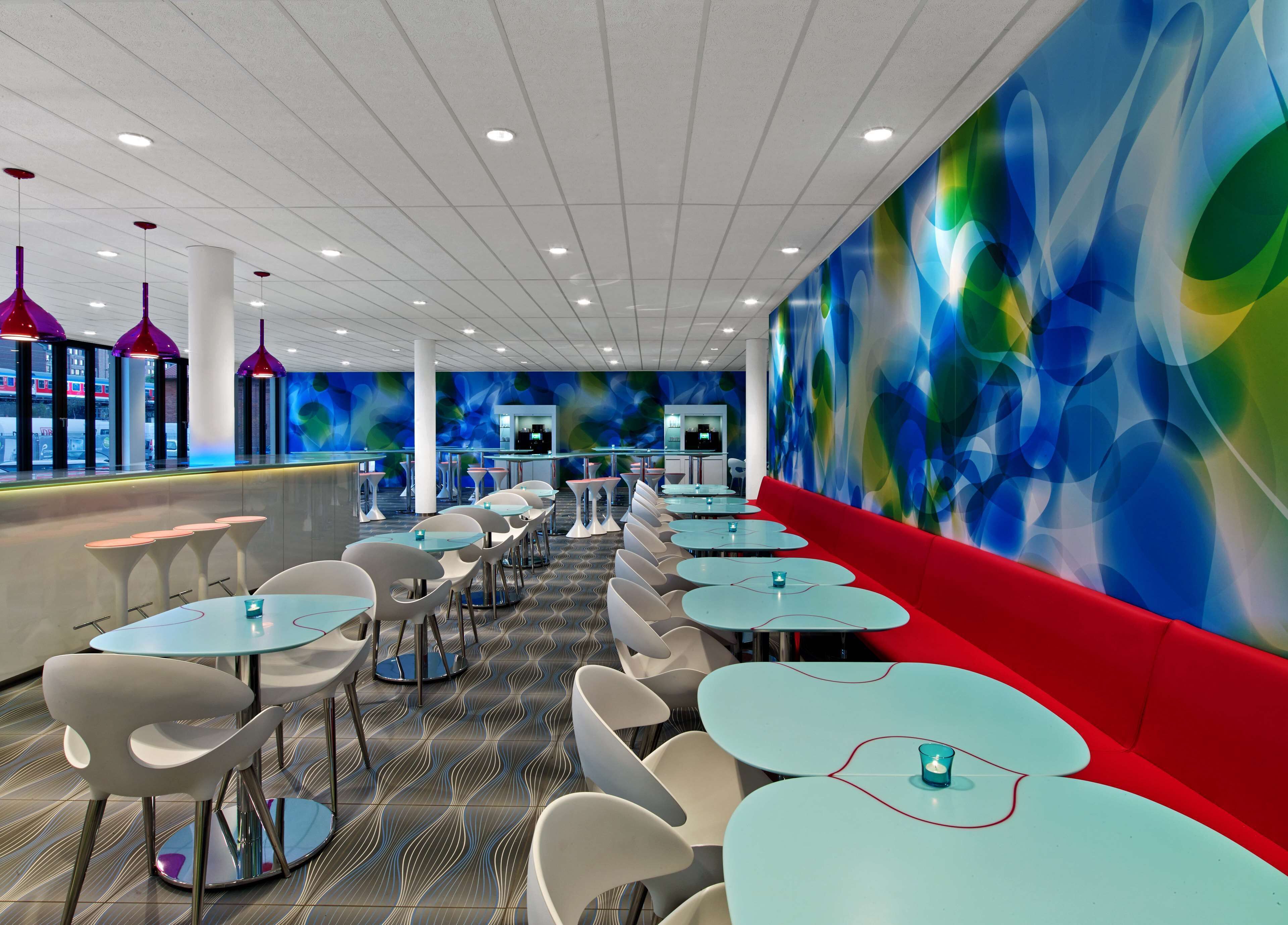 A modern restaurant with round tables and chairs, a red bench, and a colorful abstract wall.