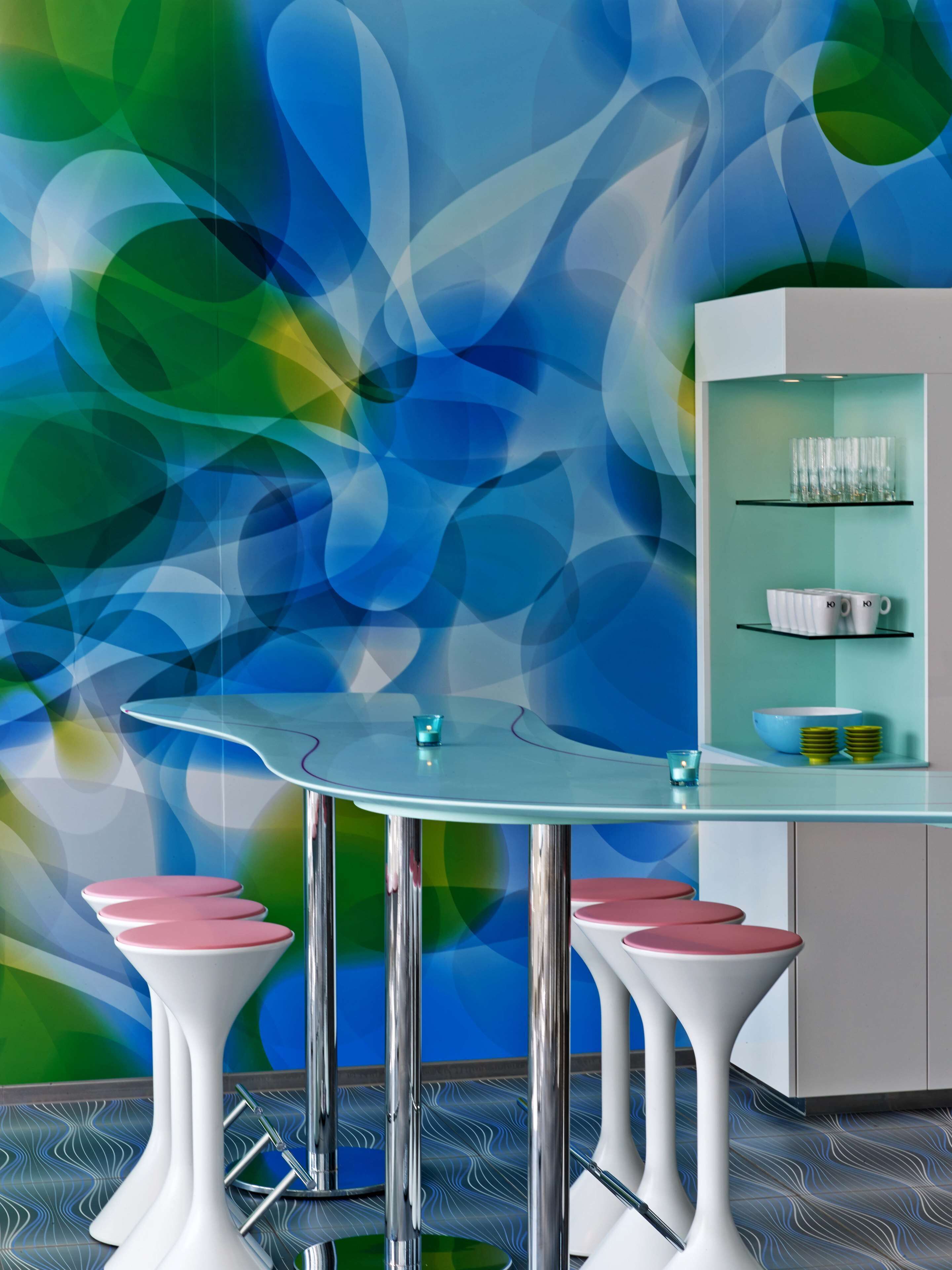 A modern bar area with a curved glass table, two pink bar stools, and a wall with abstract blue and green patterns.
