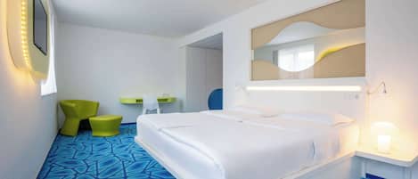 A modern hotel room with a large bed, a bedside table with a lamp, a mirror, and a seating area with a chair and ottoman.