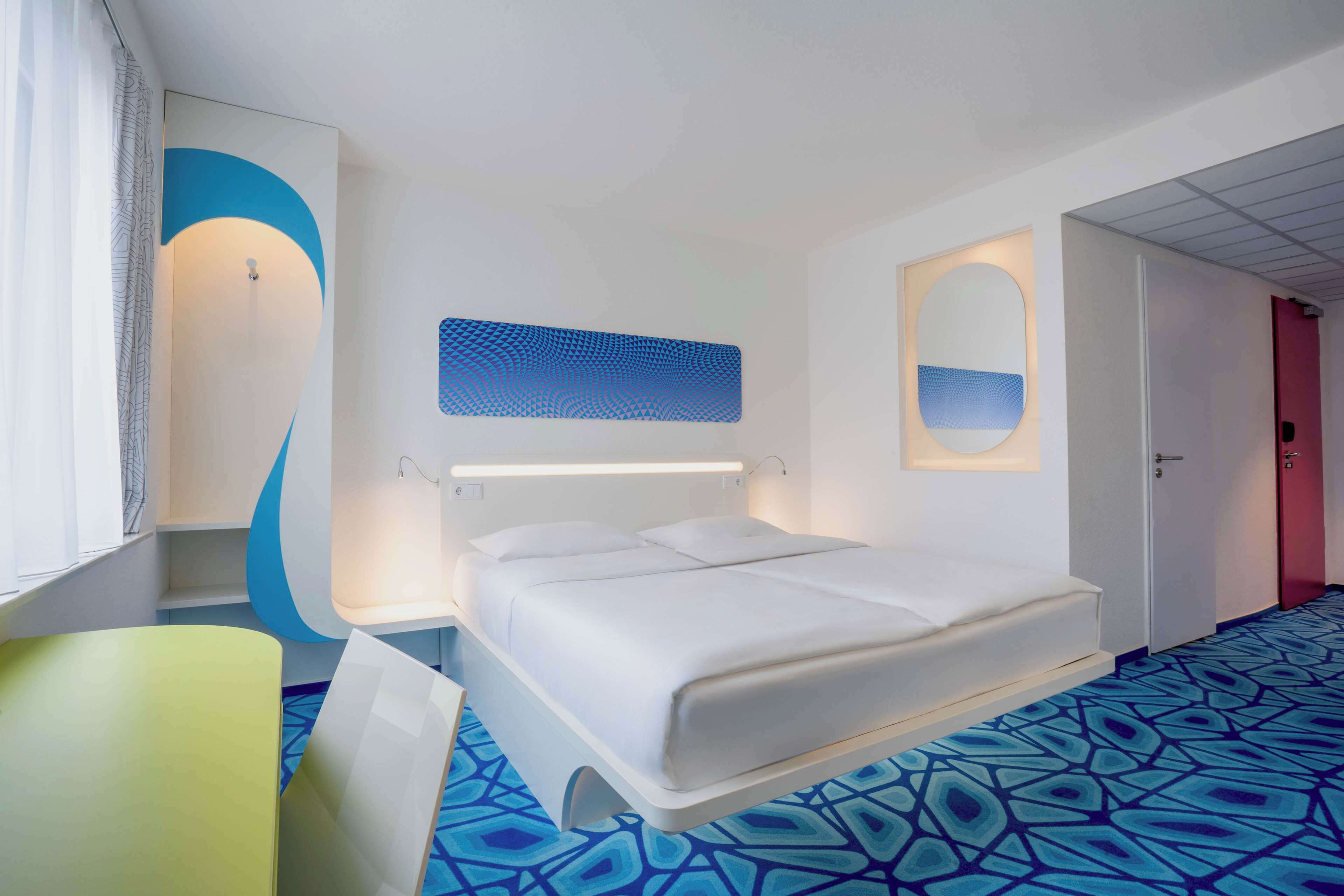A modern hotel room with a large bed, a bedside table, a chair, and a patterned floor.