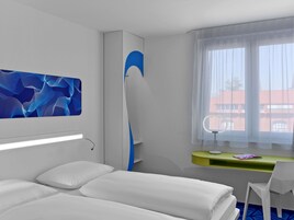 Ein modernes Hotelzimmer mit einem Bett, einem Schreibtisch mit Stuhl, einem Schminktisch mit Spiegel und einem großen Fenster mit Blick auf Gebäude.