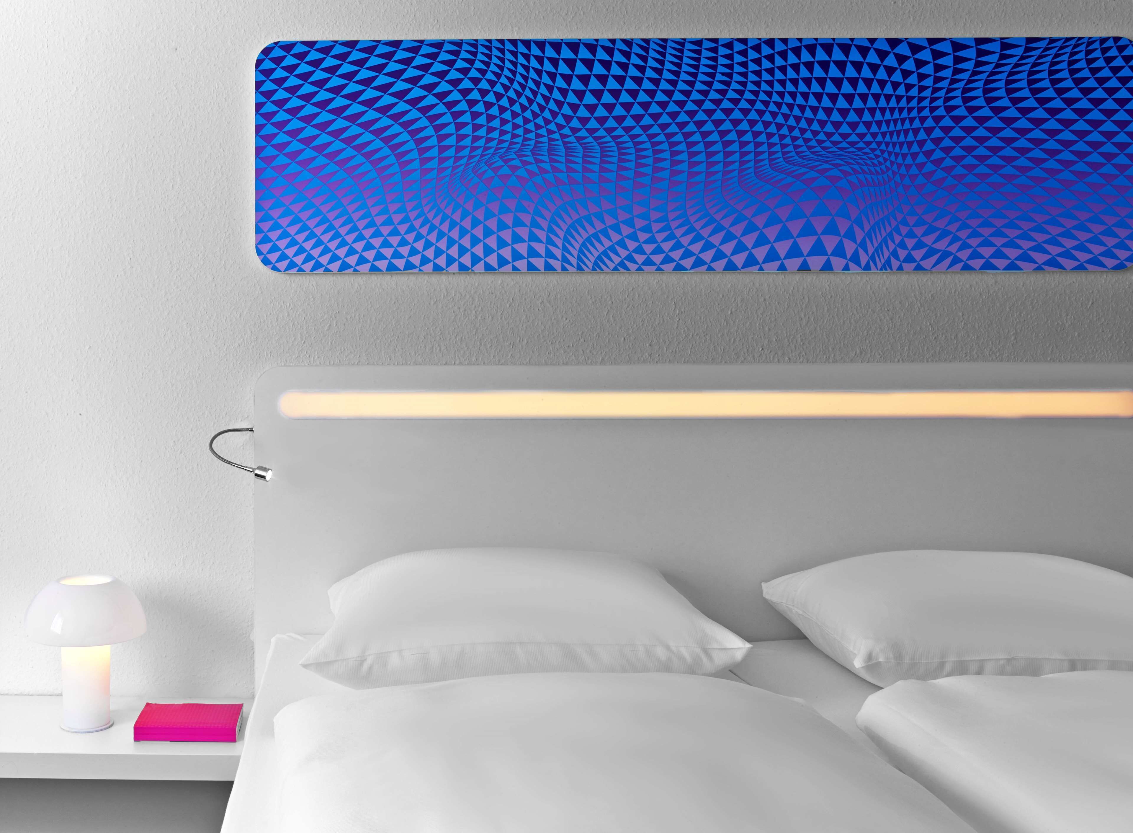A modern hotel room with a geometric-patterned headboard, a bedside table with a lamp, and neatly made beds.