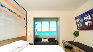A modern hotel room with a large bed, a desk with a computer, a view of the sea, and a colorful abstract painting on the wall.