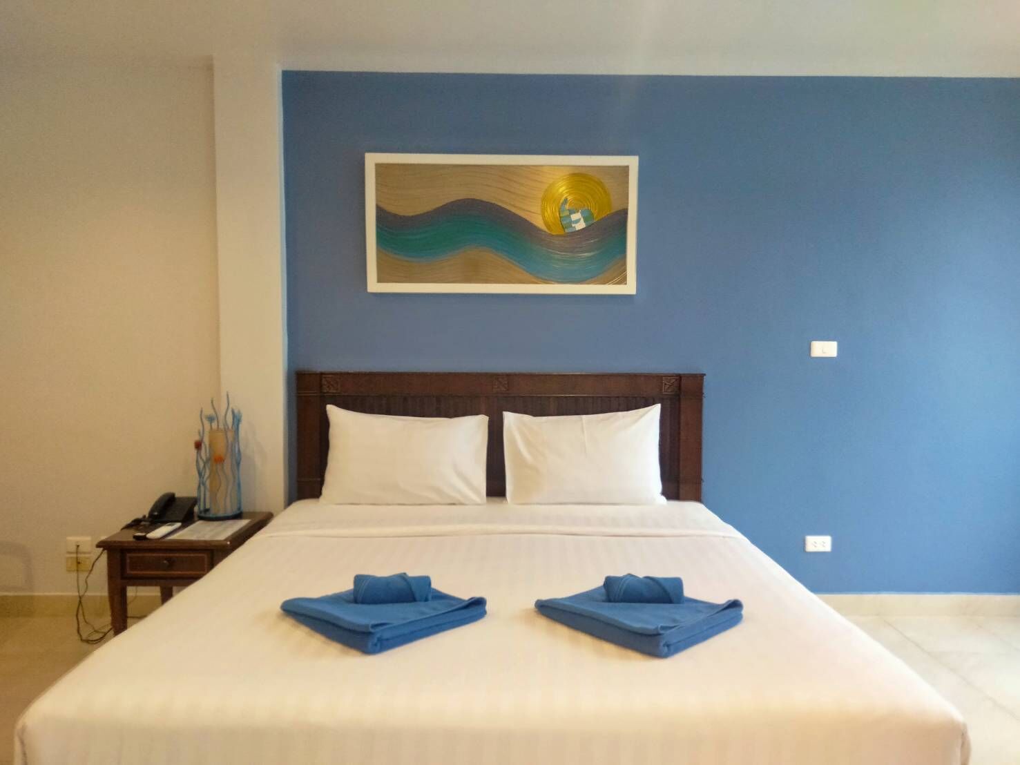 Ein Hotelzimmer mit einem großen Bett, zwei Kissen, einem Nachttisch mit Lampe, einem Bild an der Wand und zwei gefalteten Handtüchern auf dem Bett.