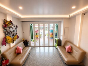 Un área de recepción con un gran mural, dos sofás, un acceso por puerta de vidrio y una pequeña mesa con una planta.