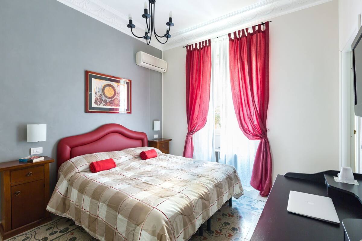 A hotel room with a bed, a desk, a chair, a lamp, a picture frame, and a window with curtains.