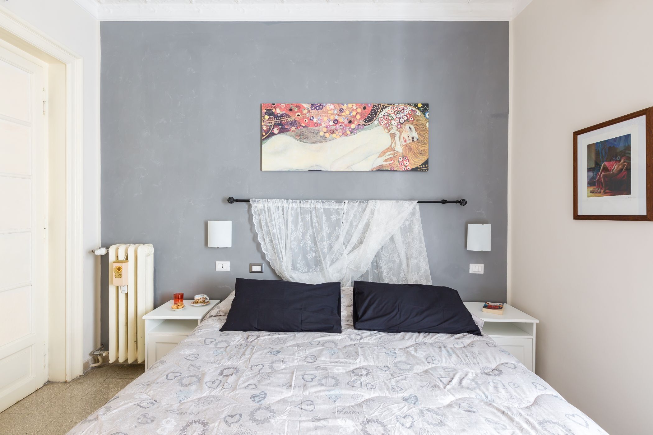 A bedroom with a bed, a bedside table, a radiator, and a wall-mounted artwork.