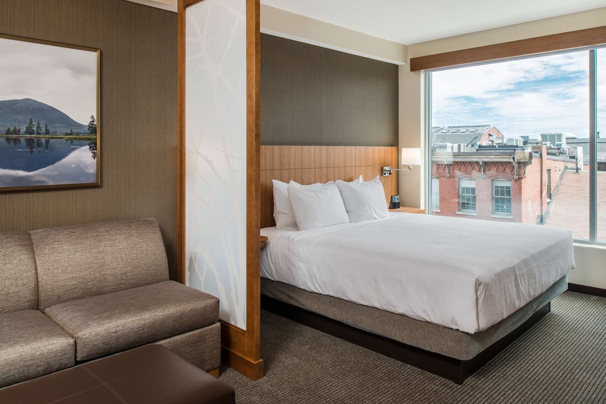 A hotel room with a bed, a sofa, a large window with a city view, and a painting on the wall.