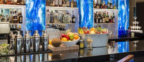A bar area with a polished granite countertop, a variety of liquor bottles, a fruit bowl, and blue upholstered bar stools.