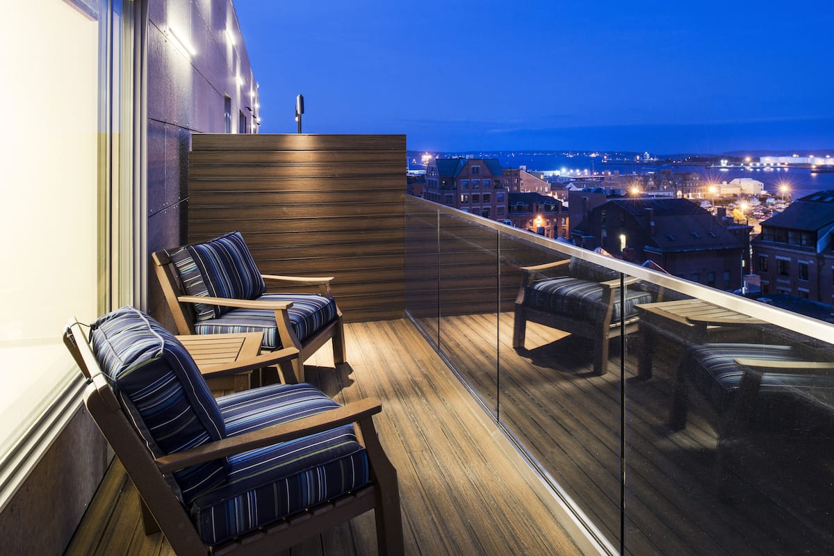 A balcony with wooden flooring, striped cushioned chairs, and a glass railing overlooking a cityscape at night.
