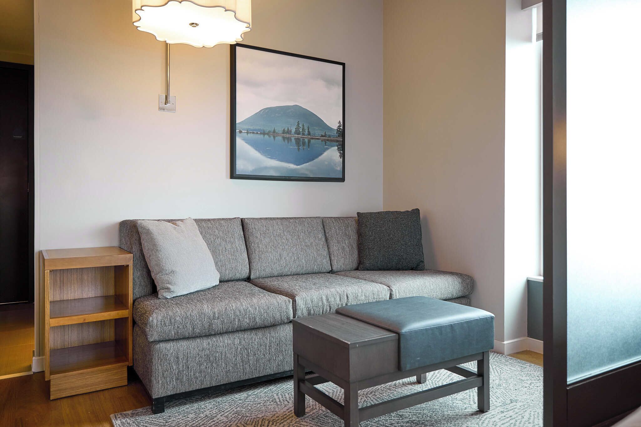 Un salon avec un canapé gris, une table d’appoint en bois, un pouf bleu et un tableau encadré représentant une montagne accroché au mur.