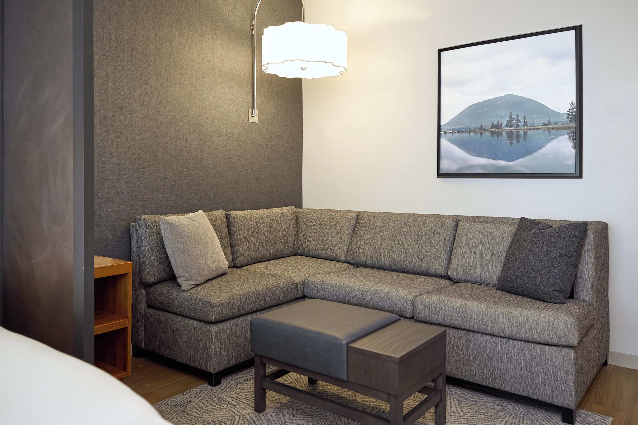 Een moderne woonkamer met een grijze hoekbank, een houten bijzettafeltje en een lijst met een landschaps schilderij aan de muur.